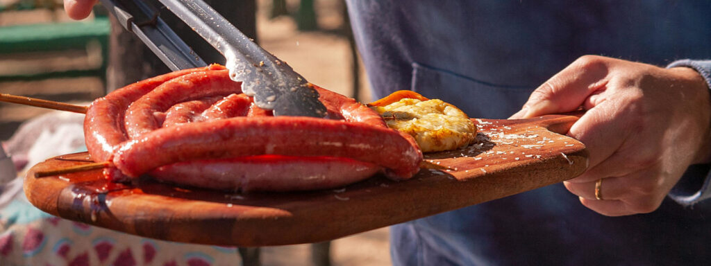 A imagem mostra um homem retirando uma linguiça da churrasqueira e colocando em uma tábua de servir